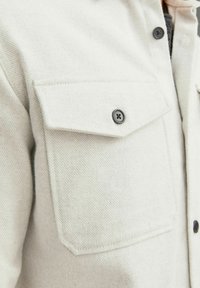 Camisa de lana de color beige claro con un acabado texturizado. Presenta dos bolsillos con solapa y botones negros en el frente y en los puños.