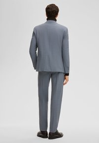 Grijs geruit colbert met bijpassende broek, slim fit, kraag plat liggend. Zwart overhemd eronder, gecombineerd met zwarte schoenen.