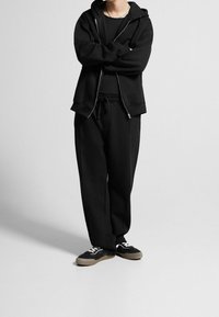 Felpa nera con zip e cappuccio, indossata sopra una maglietta nera con scollo rotondo, abbinata a pantaloni della tuta neri e larghi e a sneakers nere con lacci bianchi.