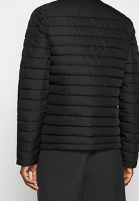 Chaquetón negro tipo puffer, con costuras acolchadas horizontales, mangas largas y una textura suave. Diseño ligero y aislante.