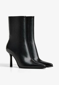 Černé lesklé kožené kotníkové boty s špičatou špičkou a úzkými vysokými podpatky na bílém pozadí.