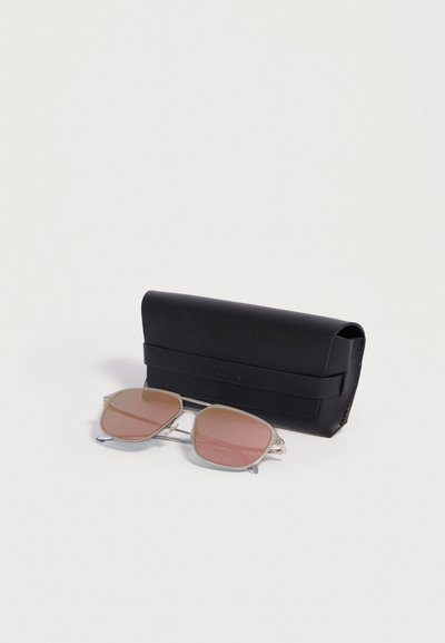 Lunettes de soleil à monture argentée avec verres teintés rose placées devant un étui en cuir noir fermé sur une surface blanche.