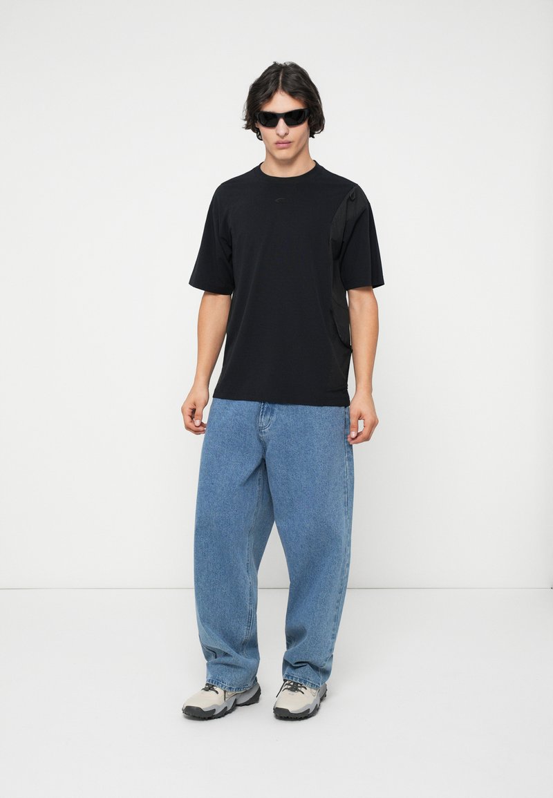 Schwarzes T-Shirt mit kurzen Ärmeln und Seitentasche, kombiniert mit lockeren hellblauen Jeans und grauen Sneakern. Modell trägt dunkle Sonnenbrille.