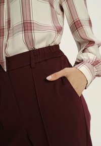 Kobieta w jasno-beżowej i burgundowej koszuli w kratę włożonej w burgundowe spodnie o wysokim stanie, z ręką w kieszeni, widoczny detal mankietu.