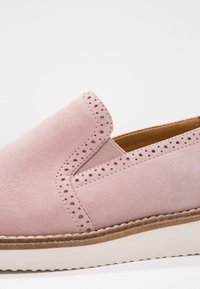 Chaussure slip-on en daim rose avec des détails perforés le long du bord, un panneau latéral élastique et une semelle en caoutchouc blanche.