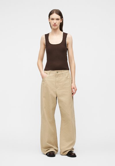 Γυναίκα που στέκεται με τα χέρια στις τσέπες, φορώντας καφέ αμάνικο τοπ με δαντέλα και μπεζ παντελόνι με φαρδιά πόδια και διακριτικές βούλες.