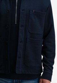 Marineblauwe button-up jas met drie grote voorkwasten over een gestructureerde marineblauwe trui met rits, gedragen met blauwe jeans.