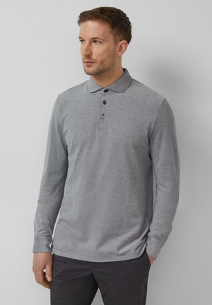 Mężczyzna ubrany w jasnoszarą koszulkę polo z długim rękawem i trzema guzikami, stojący na tle jednolitego jasnoszarego tła.