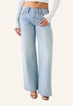 Femme portant un jean bleu clair à taille haute et jambe large, un crop top blanc côtelé et des sandales beiges, debout devant un fond uni.