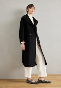 Manteau noir croisé à grand col et fentes latérales, porté sur un col roulé crème en maille côtelée et un pantalon en velours côtelé beige à jambes larges, associé à des chaussures plates grises.