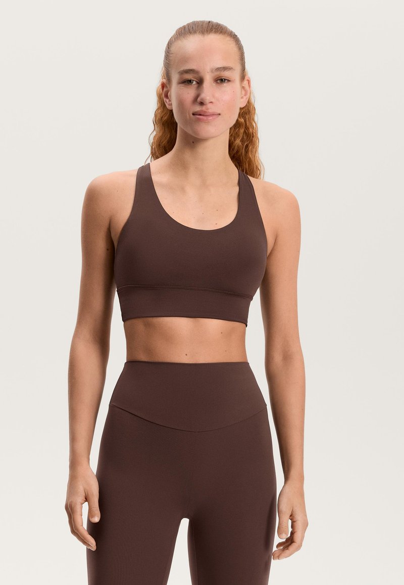 Rjavi športni modrček z zaobljenim izrezom in širokimi naramnicami, oblikovan v oprijetem slogu. Gladek teksturni videz z minimalnimi šivi.