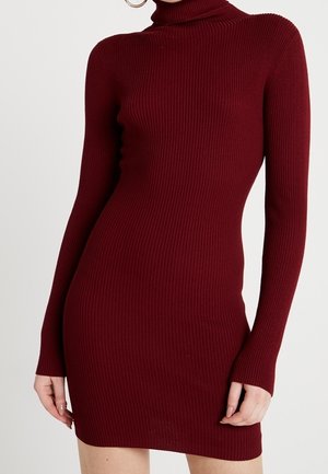 Getailleerde bordeauxrode jurk van geribbelde gebreide stof met lange mouwen en een col, gedragen door een persoon die staat tegen een effen achtergrond.