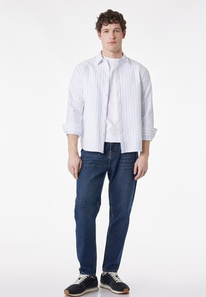 Junger Mann steht, trägt ein weiß gestreiftes Hemd über einem weißen T-Shirt, blaue Jeans und schwarze Sneakers mit weißen Schnürsenkeln.