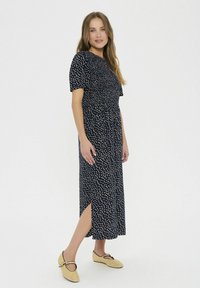 Námornícky šaty s bielymi polka bodkami, krátkymi rukávmi, priliehavým pásom a bočným rozparkom. Vyrobené z ľahkého materiálu; v súlade s béžovými, plochými šľapkami.