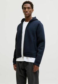 Mörkblå huvtröja med hel dragkedja, ribbade muddar och fåll, med sidofickor. Bärs över en vit t-shirt och matchas med mörka byxor.