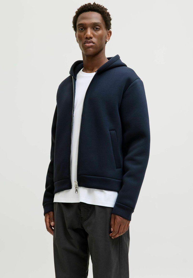 Mörkblå huvtröja med hel dragkedja, ribbade muddar och fåll, med sidofickor. Bärs över en vit t-shirt och matchas med mörka byxor.