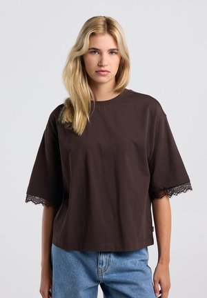 Femme blonde portant un t-shirt ample marron foncé avec des manches bordées de dentelle et un jean bleu clair, debout devant un fond uni.