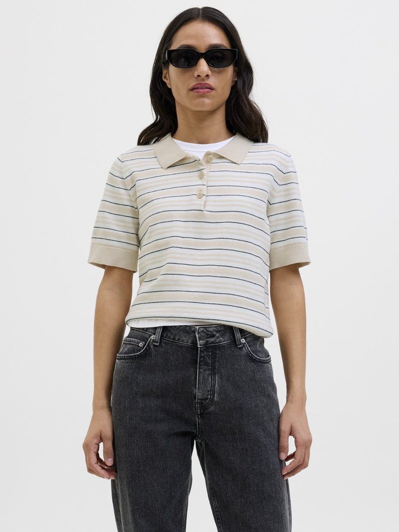 Femme portant des lunettes de soleil noires, un polo beige à manches courtes rayé, un maillot de corps blanc et un jean noir taille haute, debout devant un fond blanc.