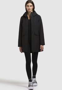 Black parka jacket with a hood, front zipper, two large front pockets, and drawstrings. Paired with black leggings and white sneakers.