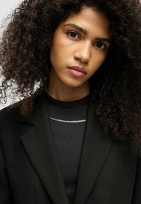Blazer nero con rever affilati, indossato su un top nero a coste con un accentuato catena d'argento; il modello ha i capelli ricci.
