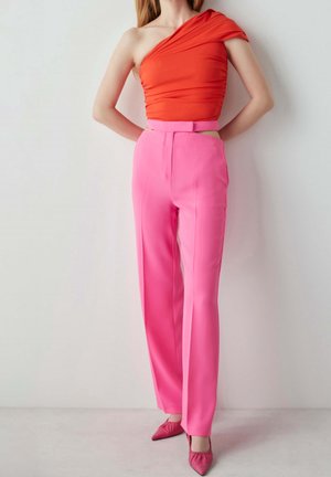 Mujer con top naranja de un solo hombro, pantalones rosa de talle alto y tacones puntiagudos rosas, de pie contra una pared blanca.