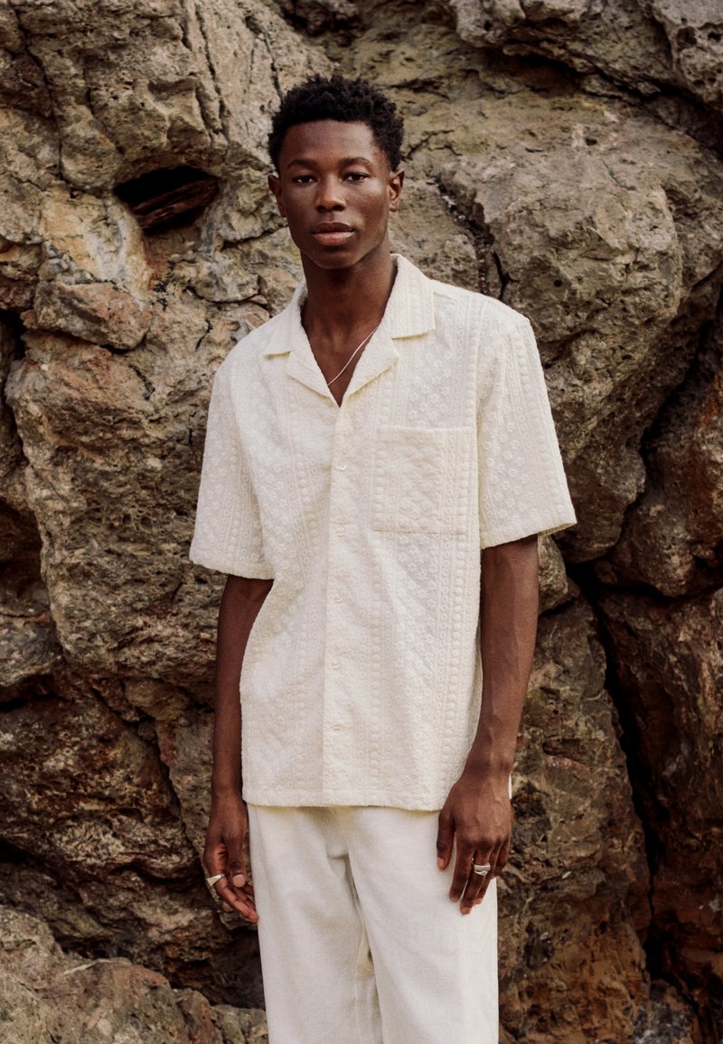 Jeune homme portant une chemise blanche texturée à manches courtes et un pantalon blanc, debout devant un fond de rochers accidentés.