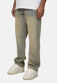 Jean en denim bleu délavé avec une coupe décontractée, présentant une jambe droite et un léger effet de frottement, associés à des baskets beiges.