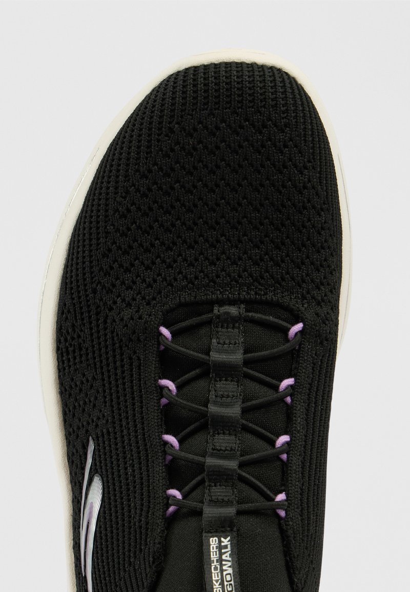 Zwarte sportieve schoen met een gestructureerde bovenkant, voorzien van paarse accenten en een vetersysteem vrij ontwerp. Witte buitenzool, lichtgewicht constructie.