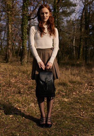 Jupe plissée marron, haut côtelé crème, sac à dos en cuir noir avec un rabat rond et des bretelles sombres, porté avec des collants et des chaussures noires. Fond forestier.
