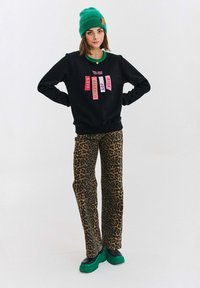 Černá mikina s barevnými textovými nášivkami, spárovaná s hnědými kalhotami s leopardím potiskem a zelenými teniskami na platformě. Zelená čepice dotváří vzhled.