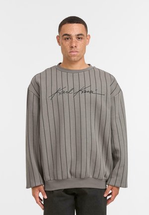 Jeune homme aux cheveux courts et aux taches de rousseur portant un pull oversized gris avec de fines rayures verticales noires et un logo distinctif sur la poitrine.