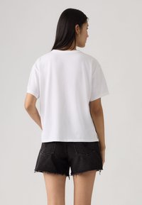 Γυναίκα με μακριά σκούρα μαλλιά, που φοράει απλό λευκό κοντομάνικο μπλουζάκι και μαύρα τζιν σορτς με ξεφτισμένα άκρα, στέκεται γυρισμένη προς τα πίσω.