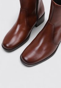 Bottes chelsea en cuir marron avec une finition brillante, dotées d'un bout arrondi, d'une fermeture éclair sur le côté et d'un petit talon carré. Texture lisse et design simple.