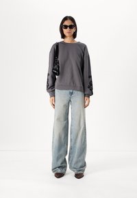 Sweatshirt gris avec des détails brodés noirs sur les manches, associé à un jean bleu délavé à large jambe. Le mannequin porte des lunettes de soleil noires et tient un petit sac.