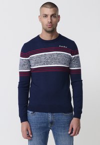 Suéter de punto azul marino con rayas horizontales en rojo, blanco y gris, que presenta un cuello redondo y puños acanalados. El logo está bordado en el pecho.