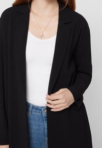 Černý dlouhý blazer s otevřeným předním dílem, nošený přes bílou halenku. Má dlouhé rukávy a volný střih, je kombinován s modrými džíny. Jednoduchý zlatý náhrdelník.