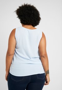 Top senza maniche di un blu chiaro, realizzato in tessuto morbido, con una sottile texture, scollatura rotonda e orlo curvo, indossato con jeans scuri.