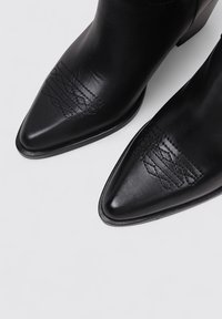 Bottes noires en cuir à chevilles avec pointes aiguës et accents de motif cousu, présentant une texture lisse et un petit talon carré.