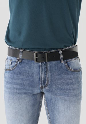 Hombre con jeans azul claro y una camisa verde azulado oscuro con un cinturón de cuero negro abrochado en la cintura.