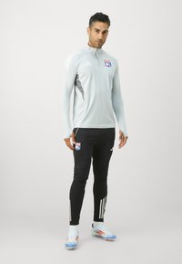 Lys grå langermet treningsgenser med halv glidelås, med OL-logoen. Svarte tettsittende bukser med tre hvite striper på sidene.