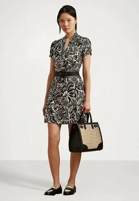 Γυναίκα που φοράει ένα φόρεμα με μαύρο και λευκό λουλούδι σχέδιο και ζώνη, κρατώντας μια μαύρη και μπεζ πλεκτή τσάντα, και φορώντας μαύρες και μπεζ λουστρίνια.