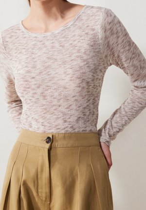 Kvinde iført en lyserød, mønstret bluse med lange ærmer, der er stoppet ned i højttaljede, vidde bukser i beige med knaplukning.