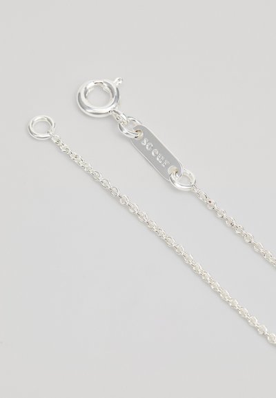 Ασημένια αλυσίδα με κούμπωμα, η οποία διαθέτει ένα ορθογώνιο κλειδί με την εγγραφή "Soeur". Η αλυσίδα έχει μια λεπτή, αλυσιδωτή σχεδίαση.