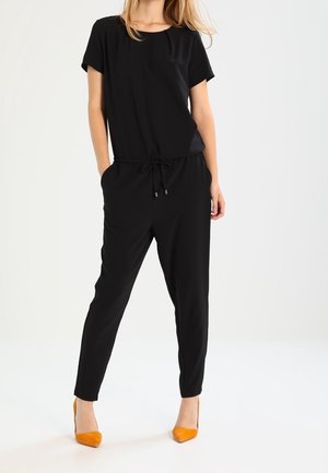 Combinaison noire à manches courtes, taille cintrée et poches latérales. Accompagnée de talons hauts orange, mettant en valeur une texture de tissu lisse.