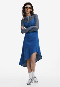 Denim mekko, jossa on epäsymmetrinen helma tummansinisessä värissä. Mekossa on istuva yläosa ja kerroksellinen muotoilu. Mallilla on päällään metallinväriset tennarit.