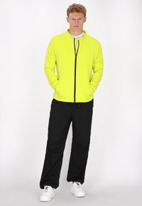 Chaqueta de cremallera amarilla neón brillante con cuello alto, combinada con pantalones negros y zapatillas blancas. La chaqueta tiene detalles de cremallera negra.