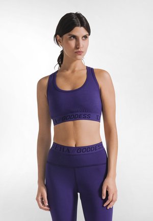 Vrouw met donker haar in een paardenstaart, gekleed in een bijpassende paarse sportbeha en legging set met de tekst "GODDESS" op de elastische banden, staand tegen een witte achtergrond.