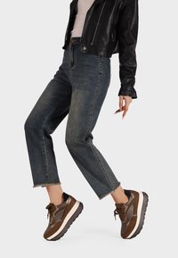 Persona che indossa jeans blu cropped, sneakers marroni e una giacca di pelle nera contro uno sfondo grigio chiaro uniforme.