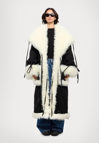 Zwarte bovenkleding met witte nepbontafwerking, grote kraag en manchetten. Voorzien van lintstrikken bij de mouwen. Draagt over jeans en dikke zwarte laarzen.