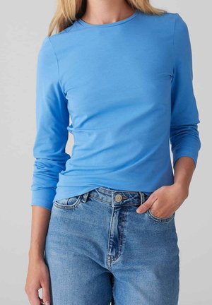 Frau trägt ein eng anliegendes hellblaues Langarmshirt, das in eine hoch taillierte blaue Jeans gesteckt ist, eine Hand in der Tasche.
